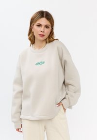 Losse beige sweatshirt met ribbels aan de cuffs, een ronde halslijn en een geborduurd groen logo op de borst. Zachte stoftextuur, minimalistisch ontwerp.