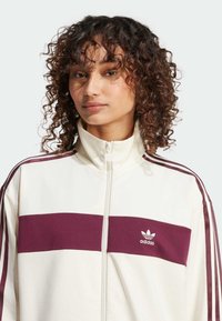 Bijela jakna na zip s bordo horizontalnom prugom, s tri bordo pruge na svakoj rukavu i izvezenim Adidas logotipom.