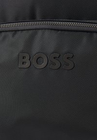 Juodas tekstūruotas audinys su metaline užtrauktuko juosta ir įspaustu BOSS logotipu centre, pabrėžiantis krepšio detalumą ir prekės ženklą.