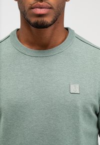 Mintgrüner Strickpullover mit Rundhalsausschnitt und gerippten Bündchen. Mit einem kleinen Stofflogo-Patch auf der linken Brust. Strukturierte Oberfläche.