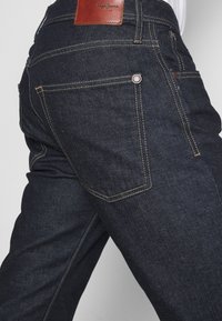 Mörkblå denimjeans med en åtsittande design, med guldstickningar, brunt läderbrandingpatch och två bakfickor med knappdetaljer.