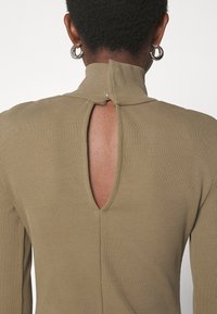 Ribbet, olivengrøn top med høj krave og en åben rygdetalje, der har en lille lukning ved halsen og et tekstureret stof.