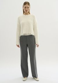 Weißer strukturierter Pullover mit einem cropped Design und fransigen Kanten, kombiniert mit grauen, gestreiften weit geschnittenen Hosen und hellen Schuhen.
