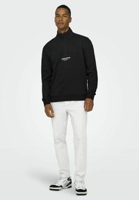 Black half-zip sweatshirt with ribbed cuffs and hem, featuring white logo text on the chest, paired with white pants and black-accented sneakers.