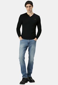 Maglietta nera a maniche lunghe con scollo a V e piccolo logo verde, abbinata a jeans azzurri e scarpe nere, in piedi con le mani in tasca.