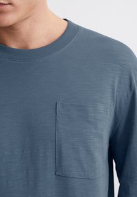 Langärmliges Hemd aus blauer Baumwolle mit strukturierter Oberfläche, Rundhalsausschnitt und einer Brusttasche. Verfügt über feine horizontale Streifen und weite Manschetten.