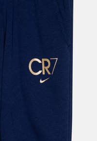 Námorné bavlnené joggery so zlatým logom CR7 a swooshom Nike. Obsahujú bočné vrecká a mäkký, textúrovaný materiál pre pohodlie.