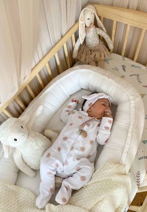 Wiege mit einer weichen, weißen Matratze. Baby in einem hellen Schlafanzug mit Sonnenmustern, umgeben von einem plüschigen Lamm und einer Stoffpuppe. Weiche Texturen sind sichtbar.