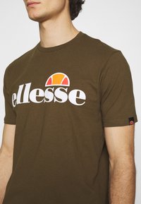 Olīvu zaļš kokvilnas t-krekls ar baltu 'ellesse' logotipu un krāsainu logotipa akcentu. Īsas piedurknes un apaļš kakla izgriezums.