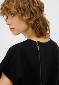 Abbigliamento nero con una vestibilità comoda, maniche corte e un zip visibile sul retro. Realizzato in materiale morbido e liscio.