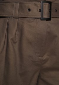 Hnědé látkové kalhoty s texturovaným povrchem, vybavené černým kovovým pásem s přezkou a viditelnými sklady, zdůrazňující strukturovaný design.