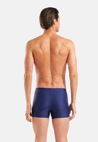 Μπλε ναυτικού χρώματος μαγιό κοντά, κατασκευασμένα από λείο, ελαστικό ύφασμα. Διαθέτουν απλό, κλασικό σχέδιο με χαμηλή μέση και χωρίς ορατά μοτίβα ή μεταλλικά στοιχεία.