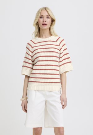 Femme blonde portant un pull en maille rayé crème et rouille avec des manches jusqu'au coude et un short blanc jusqu'aux genoux.