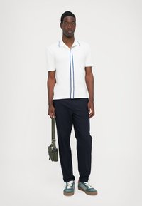 Polo shirt alb cu mânecă scurtă și detalii albastre, asortat cu pantaloni închiși la culoare și adidași verzi. Modelul ține o geantă verde.