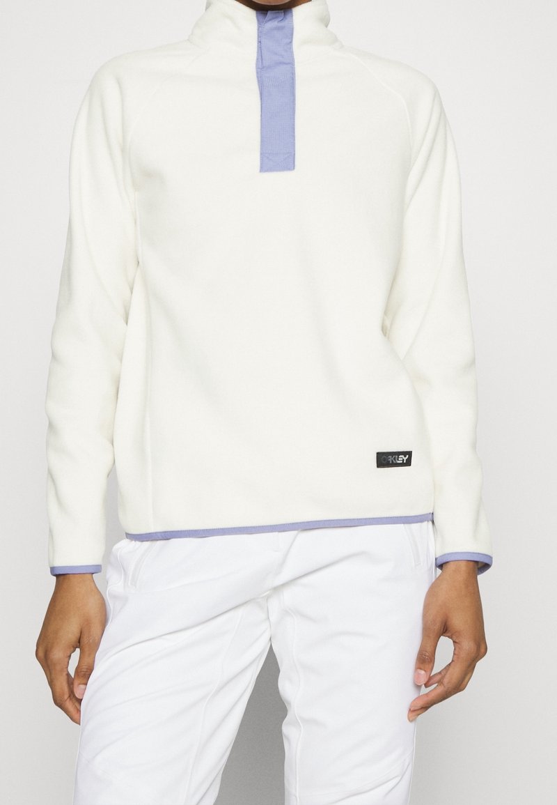 Personne portant un pull blanc avec des bordures violet clair et une fermeture éclair, associé à un pantalon blanc, debout devant un fond uni.