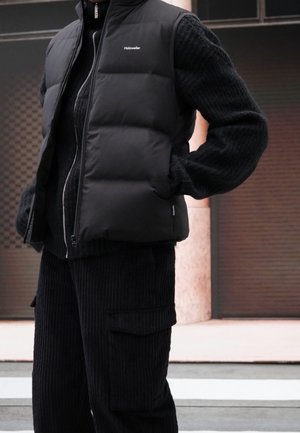Gilet rembourré noir avec col montant et fermeture éclair, superposé à un pull en tricot noir texturé. Pantalons cargo noirs doux et côtelés avec poches.