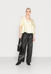 Blusa amarela de cetim com decote quadrado e mangas curtas, cintura franzida, combinada com calças de couro pretas de cintura alta e uma mala.