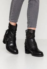 Black leather ankle boots with a pointed toe, slouchy upper, and double buckle accents, featuring a side zipper and a chunky heel.