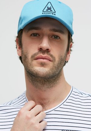 Young man with short brown hair and beard wearing a light blue cap with a black logo and a white shirt with thin navy stripes.