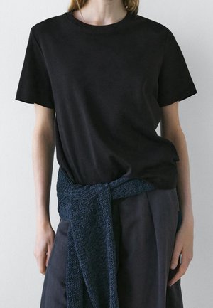 Person trägt ein schlichtes schwarzes T-Shirt mit kurzen Ärmeln und einen dunkelblauen Strickpullover, der um die Taille gebunden ist.