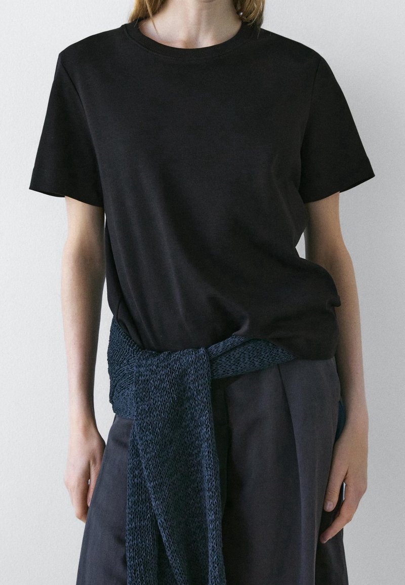 Personne portant un t-shirt noir uni à manches courtes avec un pull en tricot bleu foncé noué autour de la taille.