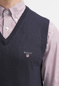 Marineblaues, gestricktes Weste mit V-Ausschnitt und GANT-Logo. Wird über einem rot-weiß gestreiften Hemd mit Kragen getragen. Weiche Textur und figurbetontes Design.