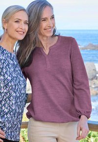 Due donne stanno all'aperto. Una indossa un top a maniche lunghe color prugna con collo henley; l'altra ha una blusa a fantasia nei toni del viola e del blu.