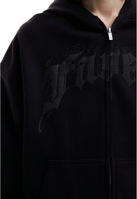 Veste à capuche noire zippée avec du texte en relief de style gothique et de petites clous décoratifs sur la poitrine, portée par une personne.