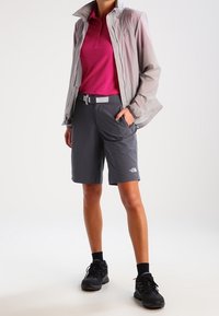 En lätt, grå jacka över en rosa pikétröja, tillsammans med mörkgrå shorts och svarta skor, med en logotyp på shortsen.
