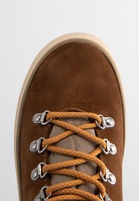 Brown suede sneaker featuring orange laces, silver hardware, and a beige rubber sole. The upper is textured and blends earthy tones.
