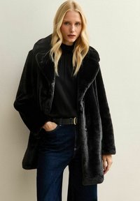 Blondynka w czarnym futrze z sztucznego materiału, czarnej bluzce i ciemnoniebieskich dżinsach, stojąca pod jasnobeżową ścianą, z ręką w kieszeni.