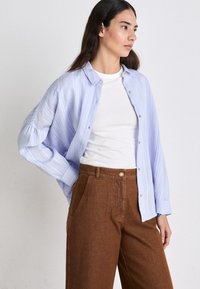 Chemise à rayures verticales bleu clair avec un design à boutons, portée sur un t-shirt blanc, associée à un pantalon large marron.