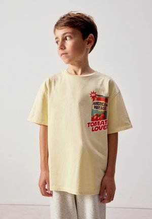 Fant, ki nosi svetlo rumeno majico s starinsko grafiko paradižnikove pločevinke v rdeči in zeleni barvi ter besedilom "Ljubitelj paradižnika", gleda vstran.