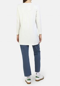 Witte tuniek-top met lange mouwen, zachte stof, afgeronde halslijn, gecombineerd met blauwe spijkerbroek met rafelige zoom en witte sneakers met groene accenten.