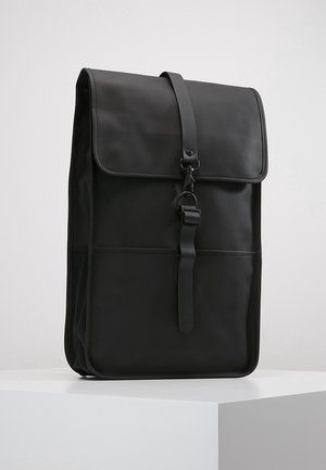 Mochila negra alta y rectangular con cierre de solapa y hebilla en la correa frontal, de pie en superficie blanca sobre fondo liso.