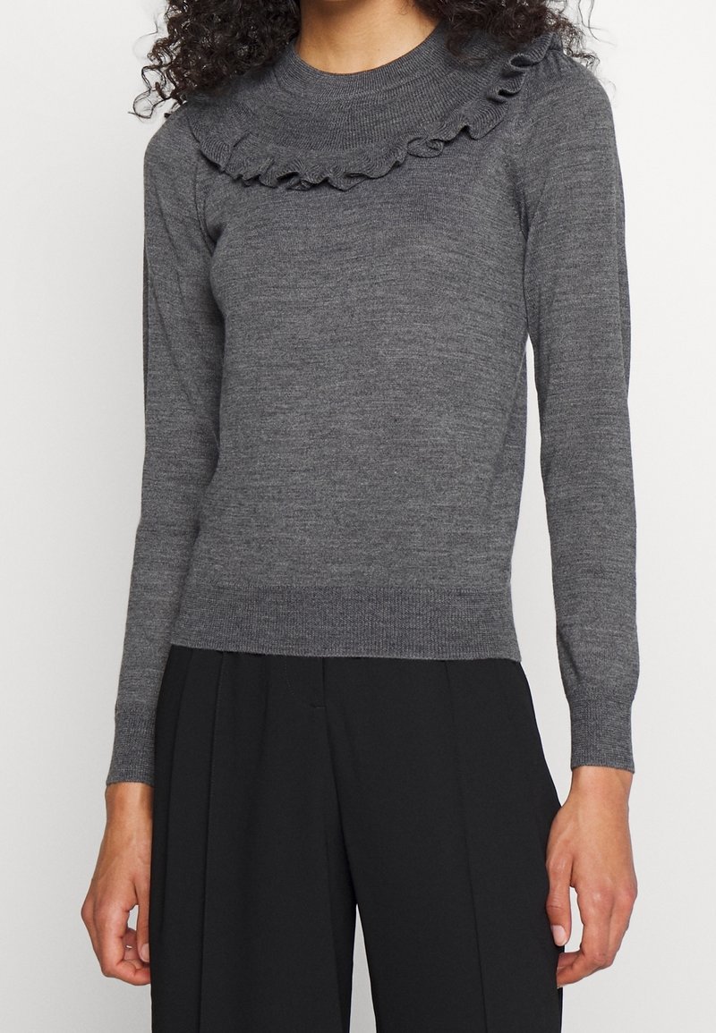 Femme portant un pull en maille gris cintré avec un détail à volants au niveau du col et des manches longues, associé à un pantalon noir.