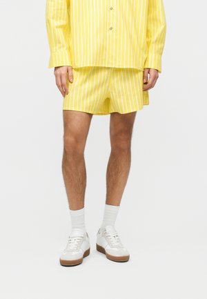 Άτομο που φοράει κίτρινες και λευκές κάθετες ριγέ σορτς και αντίστοιχο πουκάμισο, λευκές κάλτσες και λευκά αθλητικά παπούτσια με καφέ σόλες.