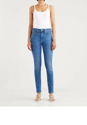 Femme portant un débardeur blanc, un jean skinny taille haute bleu et des sandales blanches à talons avec brides, debout devant un fond clair uni.