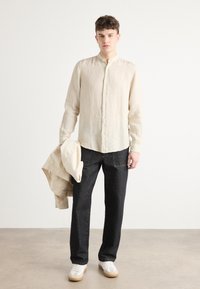 Camicia di lino beige chiaro con colletto alla coreana e chiusura a bottoni, abbinata a pantaloni in denim scuro e sneakers bianche. Tiene una giacca leggera.