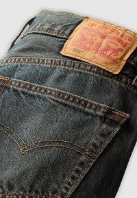 Vaqueros de mezclilla índigo con un acabado texturizado, con un parche de cuero marrón claro que presenta un logo impreso y dos bolsillos traseros con costuras naranjas.