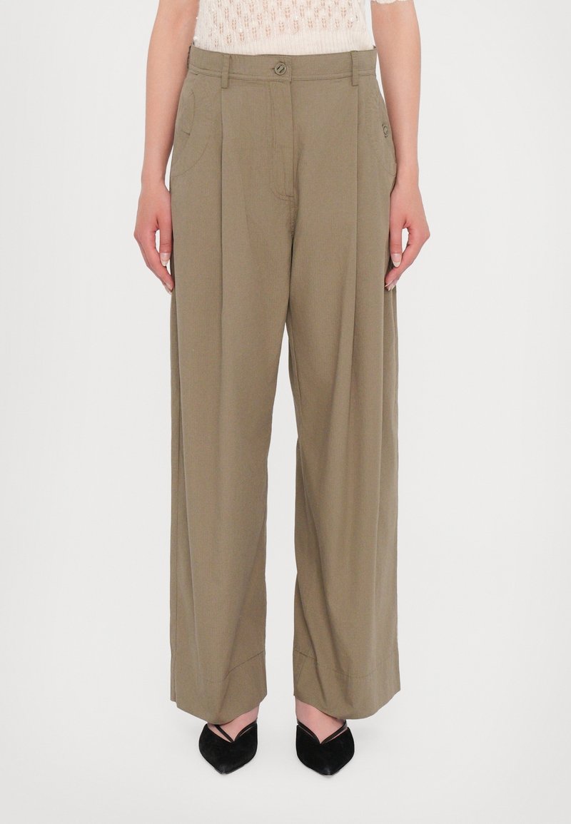 Femme portant un pantalon kaki taille haute à jambes larges et des talons noirs à bout pointu, debout devant un fond clair uni.
