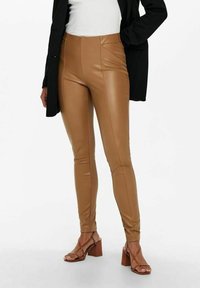 Béžové kožené legíny s vysokým pasem a přiléhavým střihem. Hladká textura s vertikálními švy, doplněné hnědými sandály na blokovém podpatku s pásky.