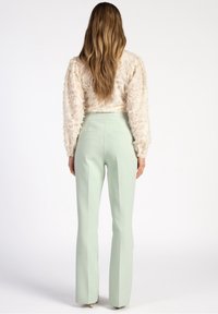 Donna con lunghi capelli mossi, che indossa una blouse cremosa a trama e pantaloni su misura verdi chiari a vita alta, in piedi contro uno sfondo bianco.