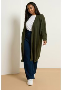 Olivgrön stickad cardigan med öppen front, långa ärmar och ribbade manschetter, ihop med högmidjade mörka jeans och vita sneakers.