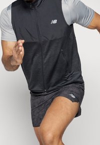 Man running in black New Balance vest, light gray short-sleeve shirt, and black patterned New Balance shorts against plain background.