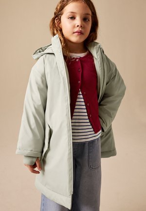 Ung pige iført en lysegrøn hættejakke, rød cardigan med knapper, stribet skjorte og denim nederdel, stående mod en beige baggrund.