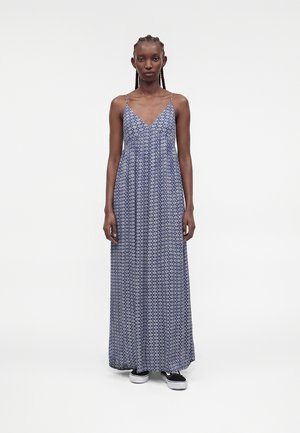 Femme aux cheveux tressés portant une robe maxi bleue à imprimé géométrique et des baskets noires sans lacets, debout sur un fond blanc uni.