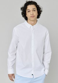 Chemise blanche à manches longues en coton, avec un col boutonné, des boutons sur le devant et un ourlet arrondi. Conception ajustée.