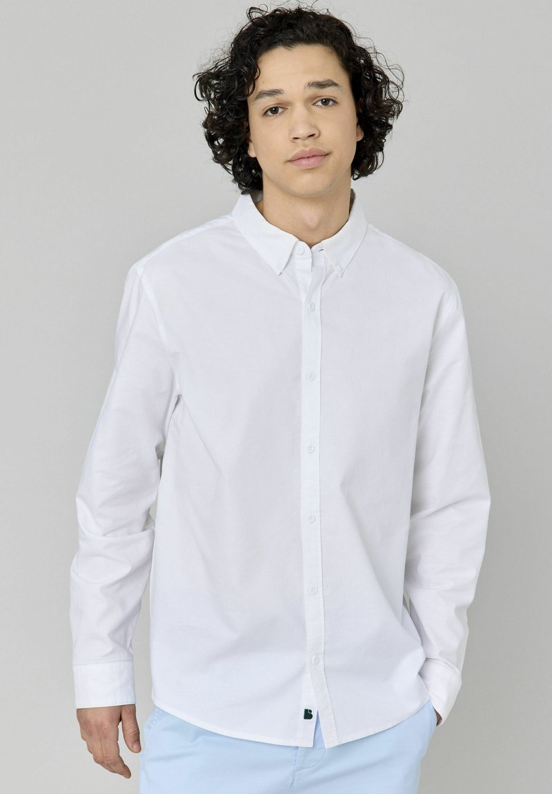 Chemise blanche à manches longues en coton, avec un col boutonné, des boutons sur le devant et un ourlet arrondi. Conception ajustée.