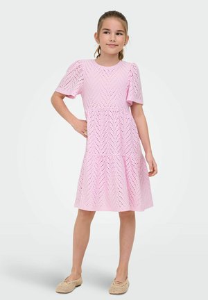 Meisje staat met hand op heup, draagt een lichtroze opengewerkte jurk met korte pofmouwen en beige geweven platte schoenen.